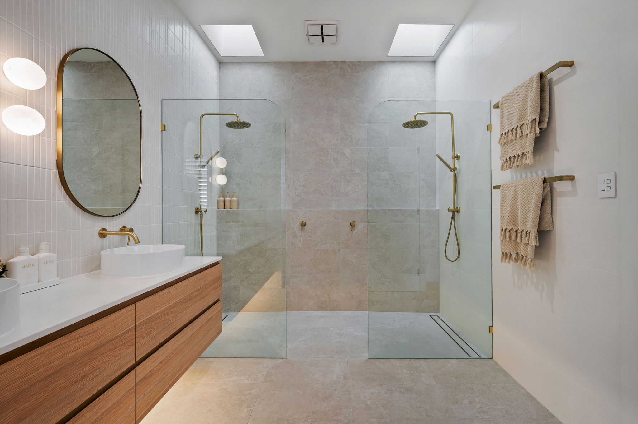 Luxury custom home bathroom design by Adelaide builders featuring dual walk-in showers with brushed gold rain shower heads, large format beige floor tiles, and a floating oak timber vanity with a white vessel basin.